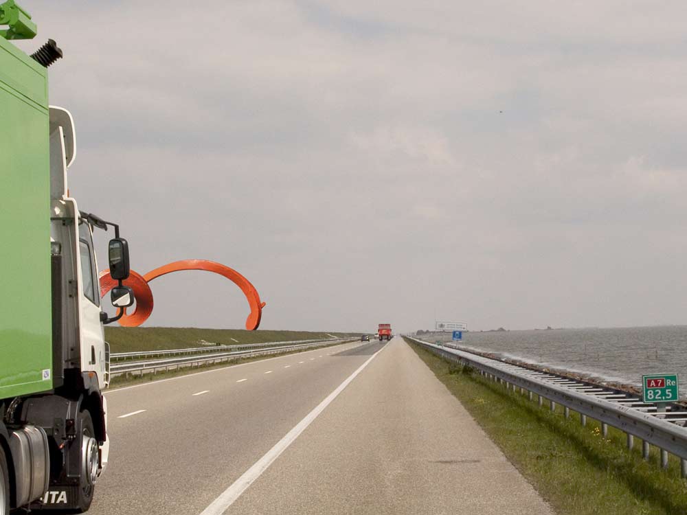 een montage, het monumentale ontwerp op de afsluitdijk verdwijnt gedeelte  achter een rijdende vrachtauto, &copy; Andr&eacute; Boone